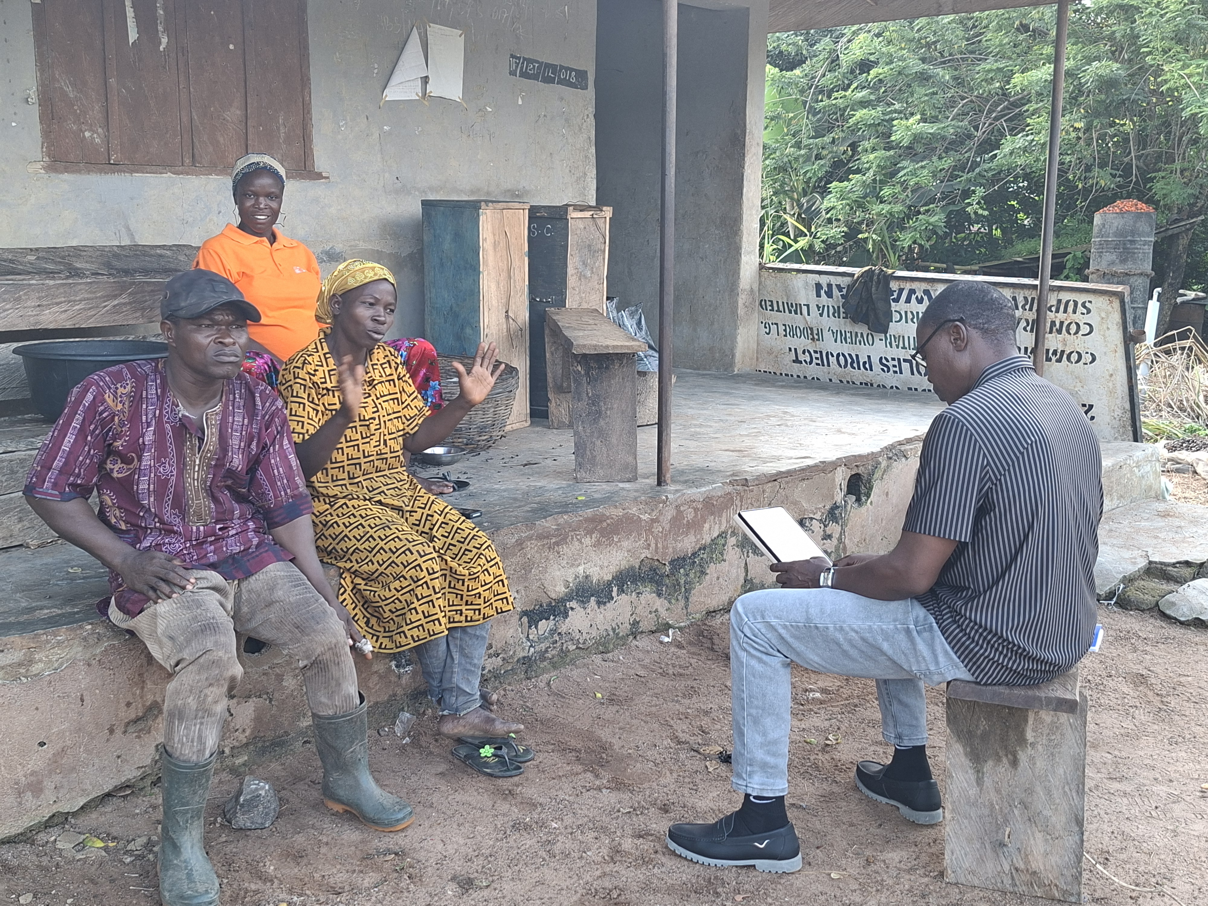 A field officer records farmers' answers on a tablet during an interview outside a rural home in Ondo State, Nigeria. Two farmers sit facing him, one speaking with hands raised.