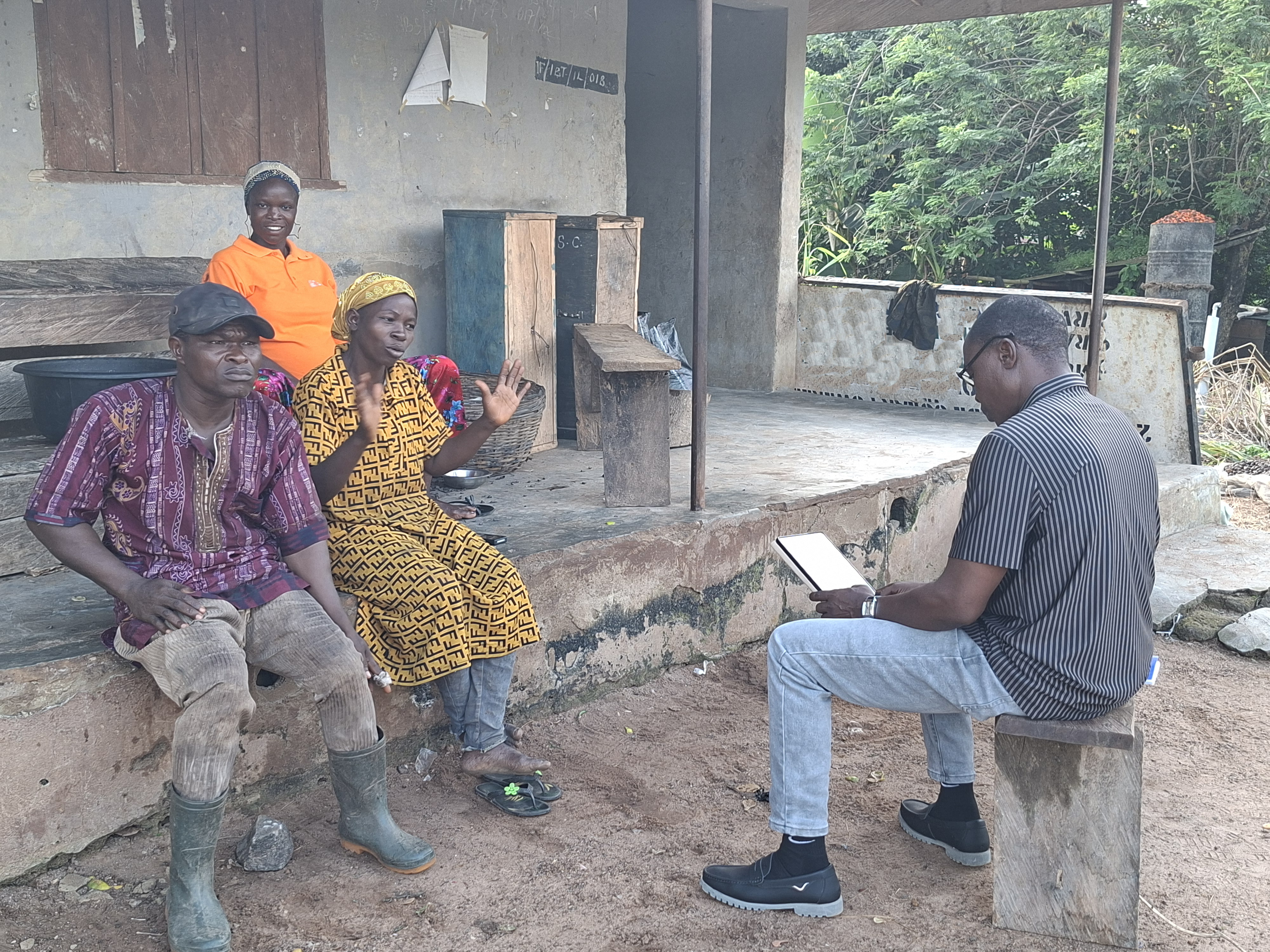 A field officer records farmers' answers on a tablet during an interview outside a rural home in Ondo State, Nigeria. Two farmers sit facing him, one speaking with hands raised.