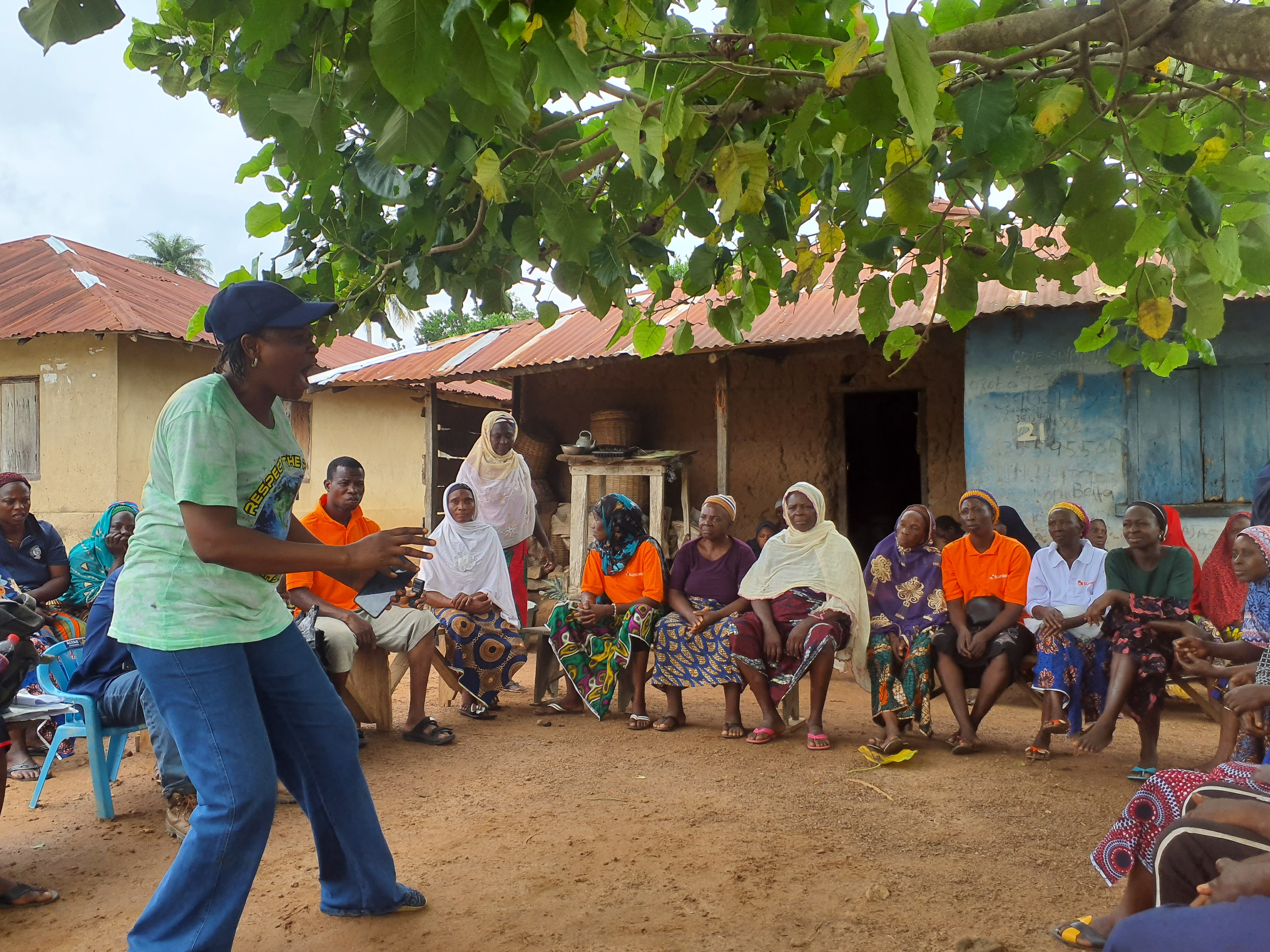 A trainer speaks animatedly to a group of women farmers seated in a circle outdoors under a tree, during a training session in a Nigerian village.