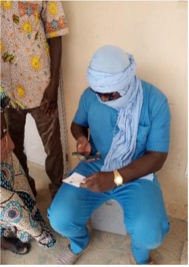 A field agent in blue scrubs and a headwrap sits outside a health facility, entering details on a phone while a caregiver and child stand beside him.