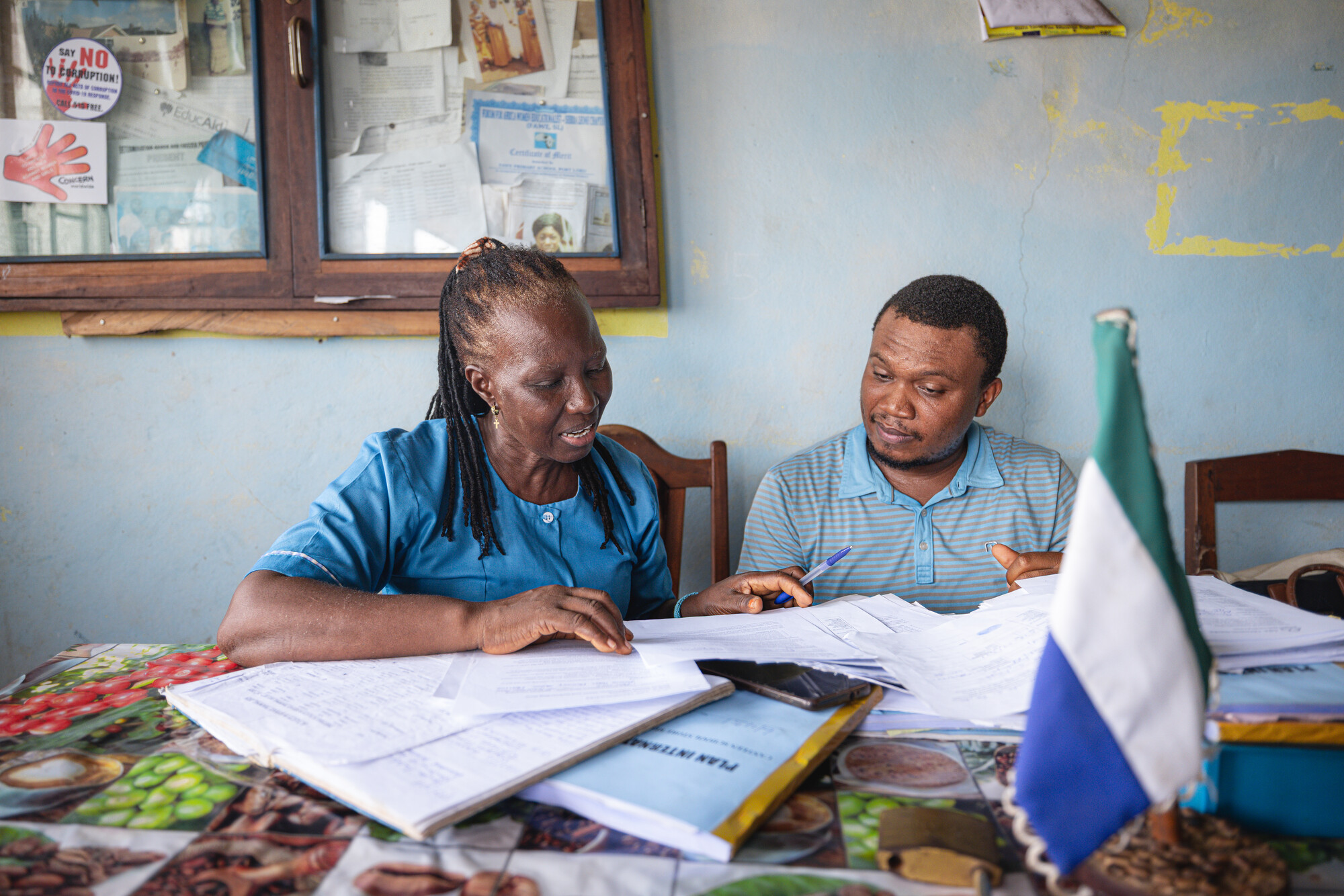 A teacher and a programme coordinator sit at a table in a school office in Port Loko, Sierra Leone, reviewing enrolment records together, with the Sierra Leone flag on the desk.