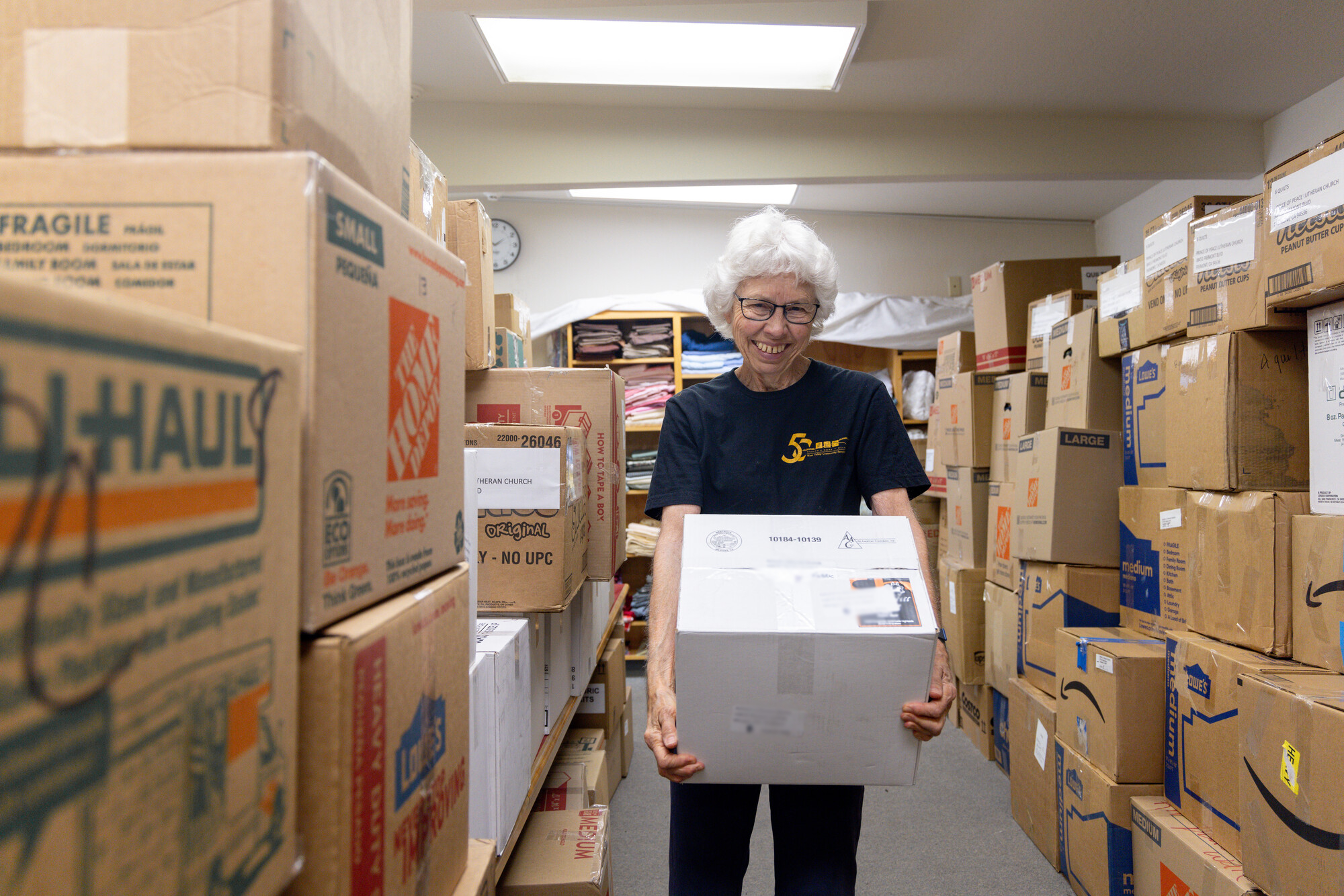 A smiling volunteer with white hair and glasses carries a labelled cardboard box down a narrow aisle stacked high with donated boxes at a church ingathering.