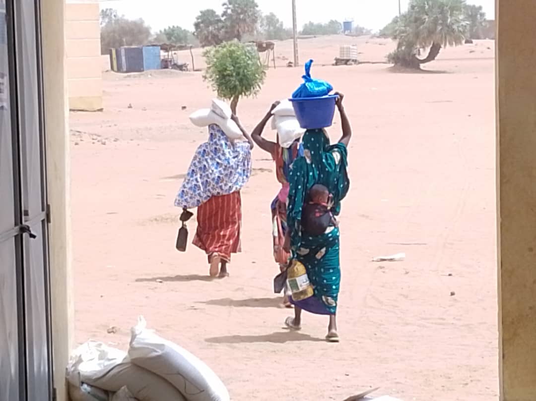Three women walking away from a health facility across open sand, carrying sacks of grain and buckets of food on their heads. One has a baby on her back.